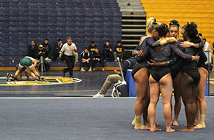 A wrestling match goes on as the Kent State gymnastics team huddles on the floor mat at the third annual Beauty and the Beast event in the M.A.C. Center on Friday, Jan. 30, 2015. The Kent State wrestling team beat Eastern Michigan University 25-9m and the Kent State gymnastics team beat the University of Pittsburgh 195.750 to 191.175.