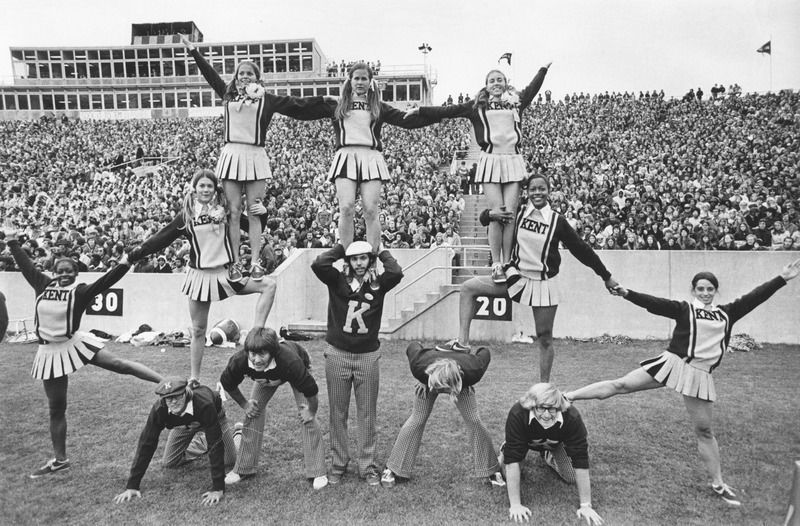 Kent State cheerleaders pose in a pyramid at a football game with the crowd in the bleachers behind them.