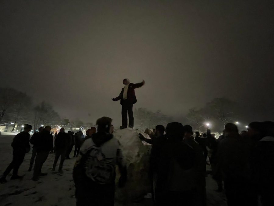 A student stands on top of giant snowball while other students attempt to roll it.