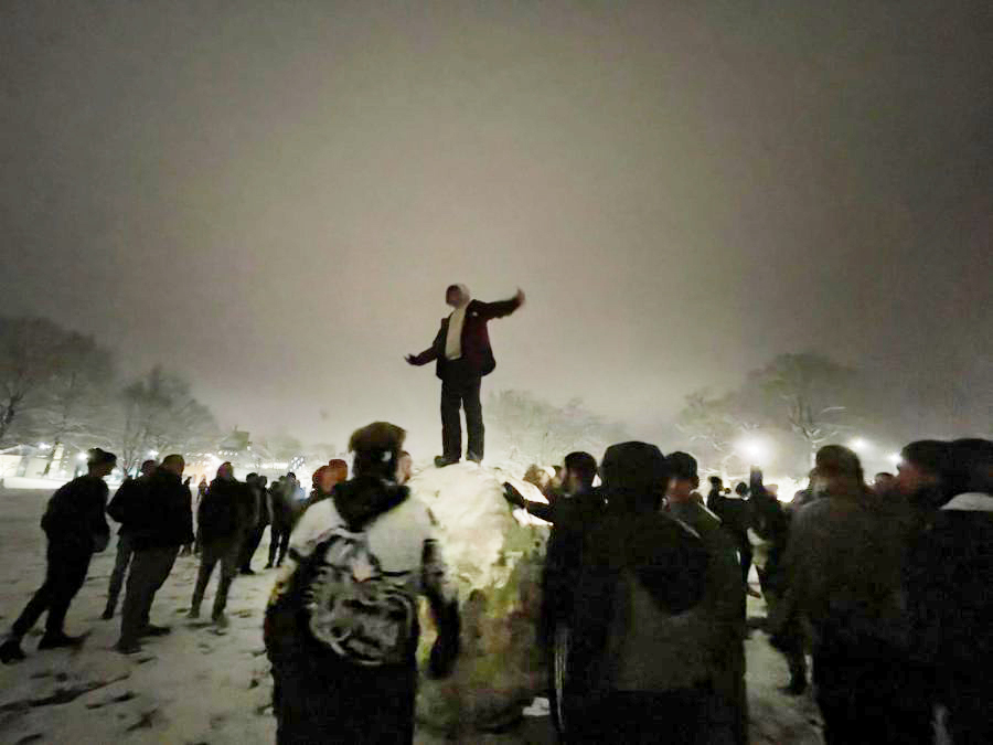 A participant of the Ski Club snowball fight rides atop the body of a giant snowball as others work to roll it across the Commons on Jan. 22, 2023.